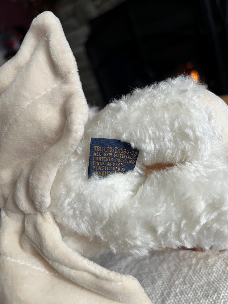 Pode incluir: Close-up de um urso de pel&uacute;cia creme. O urso tem uma textura macia e fofa e uma etiqueta com texto. A etiqueta diz "TBC LTD &copy;1990-98 ALL NEW MATERIALS CONTENTS: POLYESTER FIBER AND/OR PLASTIC BEADS HANDMADE".