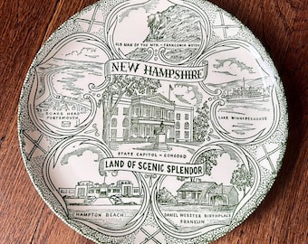 May include: A round ceramic decorative plate featuring a green and white transfer print of New Hampshire landmarks. The center displays the State Capitol in Concord, surrounded by illustrations of Old Man of the Mountain, Boars Head, Lake Winnipesaukee, Hampton Beach, and the Daniel Webster Birthplace.