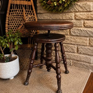 May include: A dark brown, antique wooden stool with a circular seat and turned legs. The stool is height-adjustable and rests on small caster wheels. The wood shows signs of age and wear. A small potted plant and a decorative wreath are visible in the background. The stool's height is adjustable.