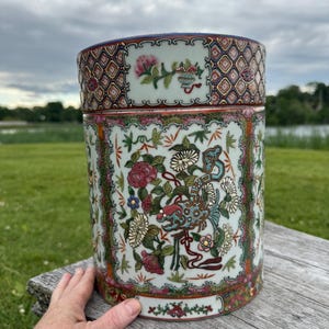May include: A cylindrical ceramic container with a lid, featuring a detailed floral and bird design in shades of red, blue, green, and white. The container is approximately 10 inches tall and rests on a wooden surface against a backdrop of green grass and a lake.