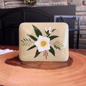 Puede incluir: Una caja rectangular de cerámica verde claro con un diseño floral pintado a mano. El diseño presenta una flor blanca con un centro amarillo, rodeada de hojas y tallos verdes. La caja se exhibe sobre una rodaja de madera.