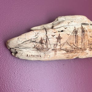 May include: Driftwood art piece with etched nautical scene. Features a lighthouse, sailboats, and flying birds. The artist's name, A. LOWTHER, is etched below the image. The driftwood is light brown against a purple background.