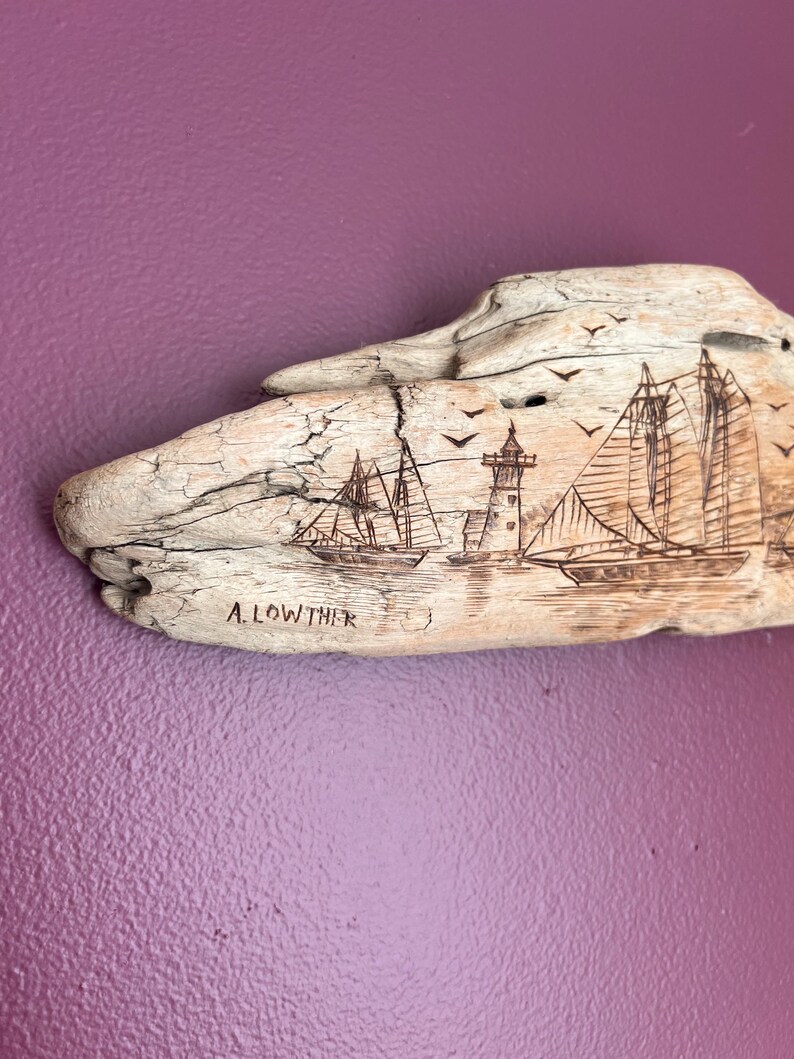 May include: Driftwood art piece with etched nautical scene. Features a lighthouse, sailboats, and flying birds. The artist's name, A. LOWTHER, is etched below the image. The driftwood is light brown against a purple background.