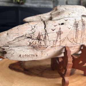 May include: A carved wooden sculpture of a ship, lighthouse, and birds. The artwork is mounted on a wooden stand. The artist's name, A. Lowther, is carved into the wood. The sculpture is made of light-colored wood.