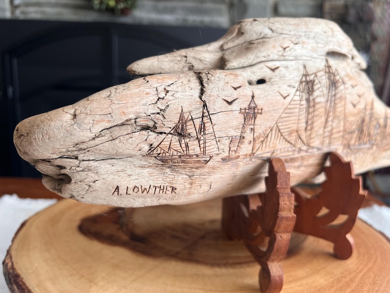 May include: A carved wooden sculpture of a ship, lighthouse, and birds. The artwork is mounted on a wooden stand. The artist's name, A. Lowther, is carved into the wood. The sculpture is made of light-colored wood.
