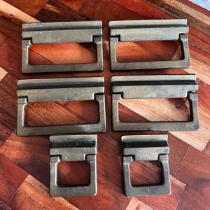 May include: Six vintage brass drawer pulls arranged on a wooden surface. The pulls have a rectangular design with a handle that folds down. The brass has a slightly aged appearance, suggesting they are antique or vintage.