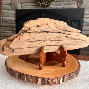 May include: A large piece of driftwood, light brown in color, is displayed on a wooden stand. The driftwood has a rough, textured surface with visible grain. The stand is made of reddish-brown wood and sits on a round wooden slice with a natural edge.