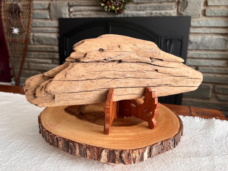 May include: A large piece of driftwood, light brown in color, is displayed on a wooden stand. The driftwood has a rough, textured surface with visible grain. The stand is made of reddish-brown wood and sits on a round wooden slice with a natural edge.