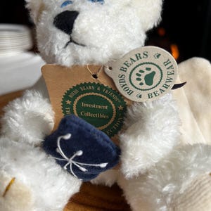 Pode incluir: Um urso de pel&uacute;cia branco com olhos azuis e nariz preto. O urso tem um chap&eacute;u azul marinho com um desenho branco e uma etiqueta que diz "Boyds Bears & Friends Bearwear Investment Collectibles". Um item de colecionador.