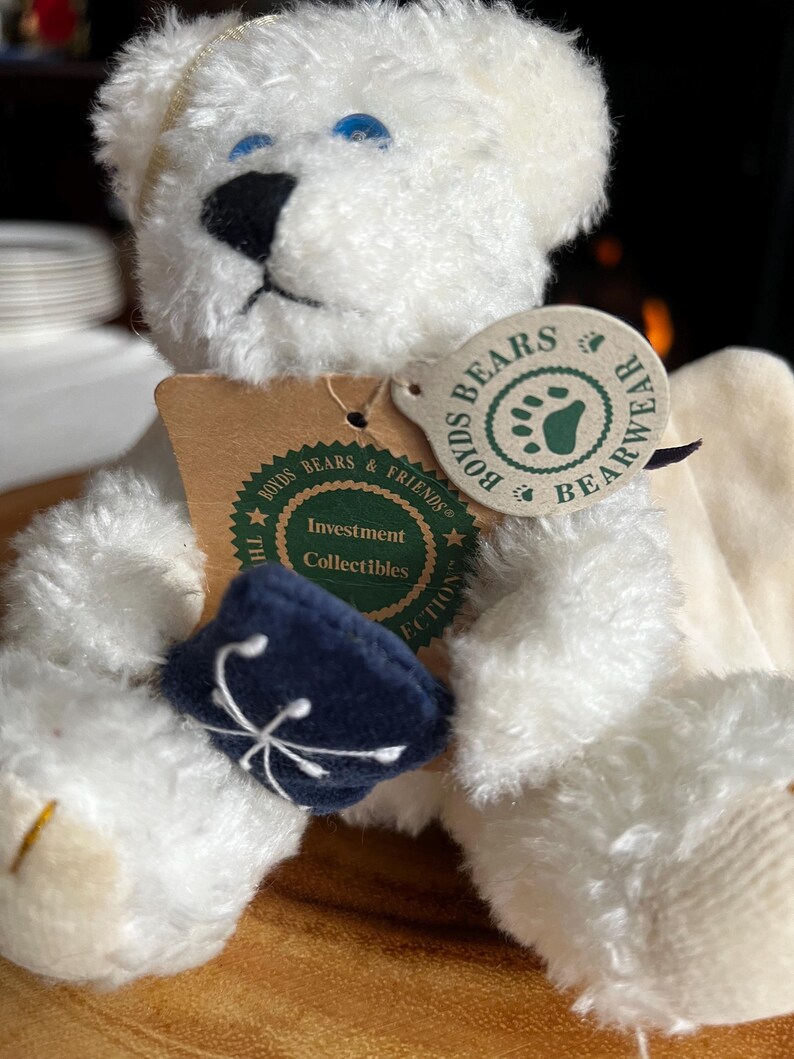 Pode incluir: Um urso de pel&uacute;cia branco com olhos azuis e nariz preto. O urso tem um chap&eacute;u azul marinho com um desenho branco e uma etiqueta que diz "Boyds Bears & Friends Bearwear Investment Collectibles". Um item de colecionador.