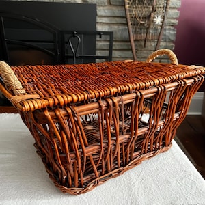 May include: A rectangular, woven wicker basket with a natural brown finish. The basket has two rope handles and is sitting on a white surface. The basket is empty and appears to be new.
