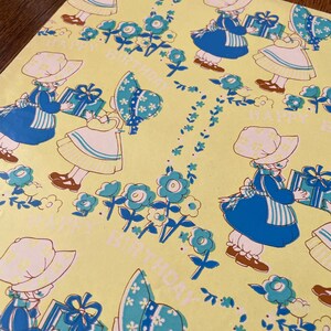 May include: Yellow wrapping paper featuring a repeating pattern of children in blue dresses and hats, holding gifts. The design includes blue flowers and the words "HAPPY BIRTHDAY".