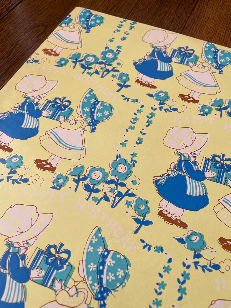 May include: Yellow wrapping paper featuring a repeating pattern of children in blue dresses and hats, holding gifts. The design includes blue flowers and the words "HAPPY BIRTHDAY".