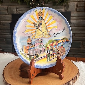May include: A decorative ceramic plate featuring a religious scene with a church, figures, and the word "Tindari". The plate has a blue border and is displayed on a wooden stand, resting on a wooden slice.