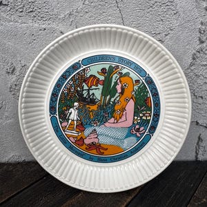 May include: White ceramic plate with a fluted rim, featuring a colorful illustration of a mermaid and a ship. The plate has text that reads "CHILDREN'S STORY 1975" and "The Little Mermaid" by Hans Christian Andersen.