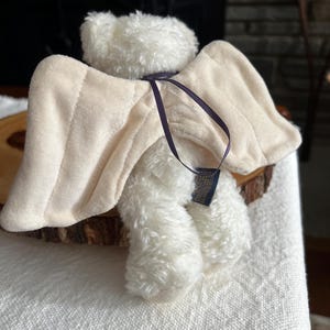 Pode incluir: Um urso de pel&uacute;cia branco com asas de anjo bege e uma fita roxa escura. As asas s&atilde;o presas &agrave;s costas do urso com uma fita roxa escura. O urso est&aacute; sentado em uma superf&iacute;cie de madeira.