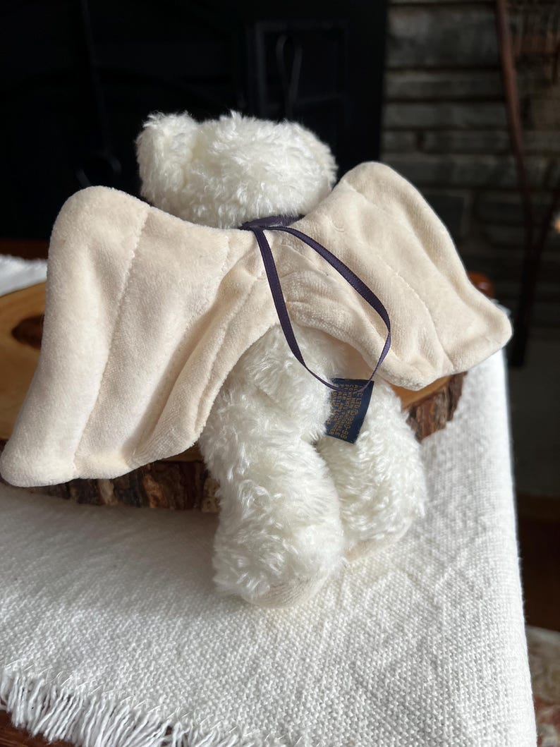 Pode incluir: Um urso de pel&uacute;cia branco com asas de anjo bege e uma fita roxa escura. As asas s&atilde;o presas &agrave;s costas do urso com uma fita roxa escura. O urso est&aacute; sentado em uma superf&iacute;cie de madeira.