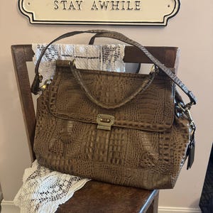 May include: A brown handbag with a textured crocodile-embossed pattern, featuring a top handle, a shoulder strap, and a metallic clasp closure. Above the bag, a white rectangular sign with black trim displays the text STAY AWHILE.