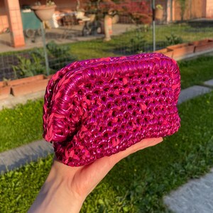 May include: A vibrant fuchsia-colored clutch with a woven design. The bag is held in a hand, showcasing its texture and color. The clutch appears to be made of a shiny material, catching the light.