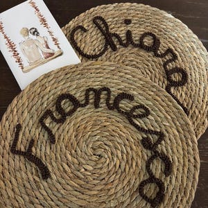 Può includere: Due sottopiatti rotondi in fibra naturale con testo marrone in corsivo "Chiara" e "Francesca". Un'illustrazione ad acquerello di una coppia su un'altalena è visibile.