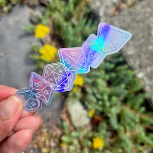 May include: A translucent, holographic sticker featuring a vertical arrangement of five polyhedral dice shapes. The design includes outlines of a d4, d6, d8, d10, and d20, each displaying its respective numerical values in a clear, thin white font.