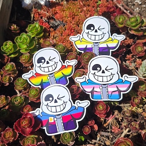 May include: Four stickers featuring a cartoon character with a white skull face, winking eye, and wide grin. Each sticker has a different jacket design with rainbow, non-binary, transgender, and other color schemes. The stickers are on a bed of green and red succulents.
