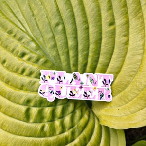 May include: A die-cut sticker featuring the text PLANT QUEER in bold, light pink block letters. The letters are decorated with small, colorful illustrations of various flowers, leaves, and vines in shades of purple, yellow, green, and black.