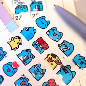 May include: A sheet of clear stickers featuring various cartoon cat-like characters in blue, yellow, and red. The stickers have different facial expressions and poses, including crying, smiling, and sticking out their tongues. A purple pen and a calendar page are in the background.