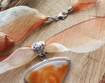May include: A pendant necklace with a large, triangular, orange agate stone set in a silver frame. The necklace is on a brown and beige ribbon.