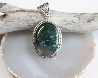 May include: A silver pendant featuring a green moss agate stone set in a silver oval frame with a decorative border. The pendant is displayed on a piece of driftwood.
