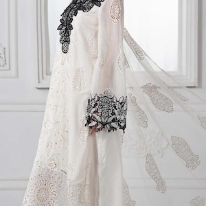 May include: White embroidered dress with black lace detailing on the neckline, sleeves, and hem. The dress has a flowing design with sheer sleeves and a matching sheer scarf. The model is wearing orange heels.