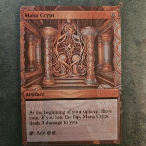 May include: A Magic: The Gathering card called "Mana Crypt". It is an artifact card with a gold and brown background. The card has an illustration of a golden crypt with pillars and intricate details. The text on the card reads: "At the beginning of your upkeep, flip a coin. If you lose the flip, Mana Crypt deals 3 damage to you. : Add 22."