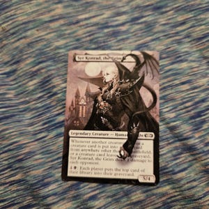 May include: A black and white Magic: The Gathering card featuring a vampire character named Syr Konrad, the Grim. The card text reads "Whenever another creature or a creature card is put into the battlefield from anywhere other than the graveyard, Syr Konrad, the Grim deals 1 damage to each opponent. 1: Each player puts the top card of their library into their graveyard." The card has a power of 5 and a toughness of 4.