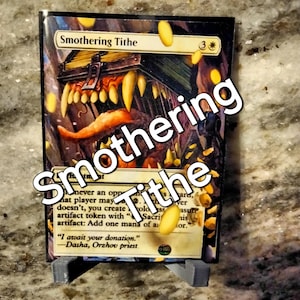 May include: A Magic: The Gathering card with a black background and a gold border. The card is titled "Smothering Tithe" and features an illustration of a monstrous creature with a large mouth and sharp teeth. The creature is holding a chest full of gold coins. The card text reads: "Whenever an opponent casts a spell, that player may pay 2 life. If they don't, you create a 1/1 colorless artifact token with "Sacrifice this artifact: Add one mana of any color."