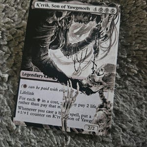 May include: A black and white Magic: The Gathering card featuring K'rrik, Son of Yawgmoth, a legendary creature with lifelink. The card text reads "(can be paid with either life or 2 life) Lifelink. For each  in a cost, rather than pay that mana, you may pay 2 life. Whenever you cast a black spell, put a +1/+1 counter on K'rrik, Son of Yawgmoth."