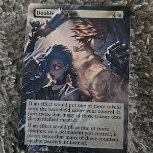 May include: A Magic: The Gathering card with a green mana symbol in the upper right corner. The card is titled "Double Down" and features an illustration of two figures with a futuristic aesthetic. The card text reads: "If an effect would put one or more tokens onto the battlefield under your control, it puts twice that many of those tokens onto the battlefield instead. If an effect would place one or more counters on a permanent you control, it places twice that many of those counters on that permanent instead."