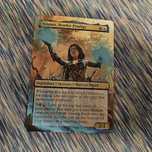 May include: A Magic: The Gathering card featuring Kinnan, Bonder Prodigy, a Human Druid creature card with a blue, green, and gold mana cost. The card art depicts a woman with dark hair and a blue and gold outfit, holding a staff and standing in front of a swirling blue and gold background. The card text reads: "Whenever you tap a nonland permanent for mana, add one mana of any type that permanent produced. 5GG: Look at the top five cards of your library. You may put a non-Human creature card from among them onto the battlefield. Put the rest on the bottom of your library in a random order. 2/2"