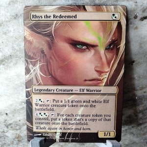 May include: A Magic: The Gathering card featuring Rhys the Redeemed, a legendary Elf Warrior creature with green and white colors. The card text reads "2, : Put a 1/1 green and white Elf Warrior creature token onto the battlefield. 4 : For each creature token you control, put a token that's a copy of that creature onto the battlefield. Whole again in honor and horn."