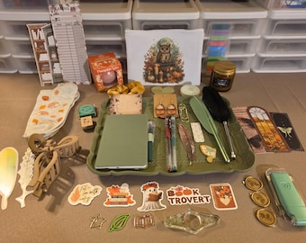 May include: A collection of stationery and autumn-themed items including a green tray, a notebook, pens, stickers, bookmarks, a pouch with an owl illustration, a candle, a pumpkin spice item, hair accessories, and wax seals arranged on a flat surface.