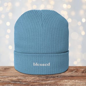 May include: A light blue ribbed knit beanie with the word "blessed" embroidered in white on the folded brim. The hat is made of a soft material and is suitable for cold weather. The background is blurred with bokeh lights.