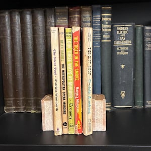 May include: A row of five vintage paperback books standing upright between two light-colored stone bookends. The titles include The Silent Pool, The Metropolitan Opera Murders, Look Three Ways at Murder, The Tiger in the Smoke, and The Gift Shop.