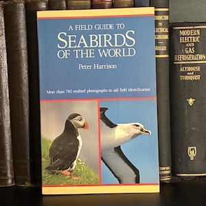 May include: A Field Guide to Seabirds of the World by Peter Harrison. The book features a blue cover with the title in white and a photo of a puffin and a seabird. The book is surrounded by other books on a shelf.
