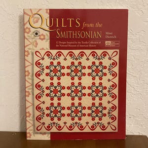 May include: Book cover featuring a red, white, and gray quilt pattern with the title "Quilts from the Smithsonian" and the author "Mimi Dietrich".