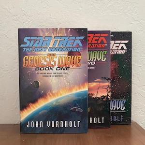 May include: Three paperback books in a row, all Star Trek: The Next Generation novels. The first book is titled "The Genesis Wave, Book One" and features a red and orange planet with a spaceship in the background. The second book is titled "The Wave, Two" and features a sunset over a forest. The third book is titled "The Wave" and features a dark, starry sky.