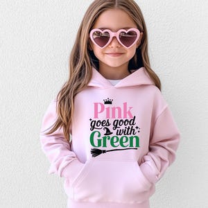 May include: A light pink hooded sweatshirt with the text "Pink goes good with Green" in pink and green. The design includes a witch hat, stars, and a broom. The sweatshirt has a front pocket and is worn by a person wearing heart-shaped sunglasses.