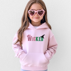 May include: A light pink hooded sweatshirt with the word "Wicked" in glittery pink and green letters, and a black witch hat graphic. The sweatshirt has a front pocket and is being worn by a young person wearing heart-shaped sunglasses.