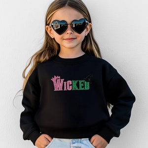 May include: Black sweatshirt with the word "Wicked" in pink and green glitter lettering. A crown graphic is above the word "Wicked". The sweatshirt is worn by a young person wearing heart-shaped sunglasses.