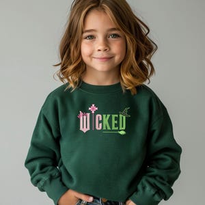 May include: A dark green sweatshirt with the word 'WICKED' printed on the front in pink and green letters. The word is surrounded by small pink and green graphic elements, including a crown, a wand, a witch's hat, and a broom.