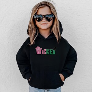 May include: Black hooded sweatshirt with the word "Wicked" in pink and green glitter lettering. The sweatshirt features a crown graphic above the word "Wicked" and a bat graphic to the right. The child is wearing heart-shaped sunglasses.
