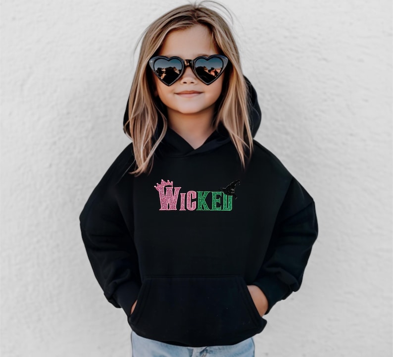May include: Black hooded sweatshirt with the word "Wicked" in pink and green glitter lettering. The sweatshirt features a crown graphic above the word "Wicked" and a bat graphic to the right. The child is wearing heart-shaped sunglasses.